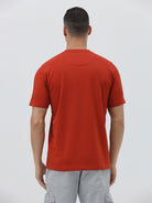 A man wearing a short-sleeve crewneck t-shirt in a muted brick red color, paired with light gray cargo pants, standing against a light gray studio background.