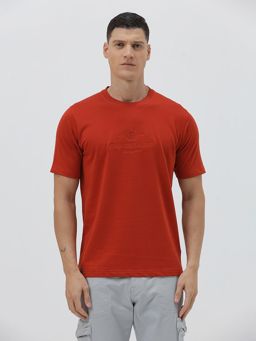A man wearing a short-sleeve crewneck t-shirt in a muted brick red color, paired with light gray cargo pants, standing against a light gray studio background.