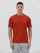 A man wearing a short-sleeve crewneck t-shirt in a muted brick red color, paired with light gray cargo pants, standing against a light gray studio background.