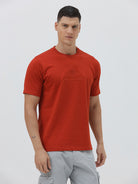 A man wearing a short-sleeve crewneck t-shirt in a muted brick red color, paired with light gray cargo pants, standing against a light gray studio background.