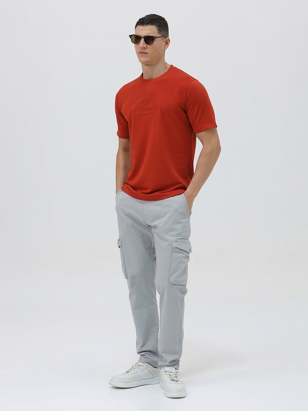A man wearing a short-sleeve crewneck t-shirt in a muted brick red color, paired with light gray cargo pants, standing against a light gray studio background.