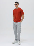 A man wearing a short-sleeve crewneck t-shirt in a muted brick red color, paired with light gray cargo pants, standing against a light gray studio background.