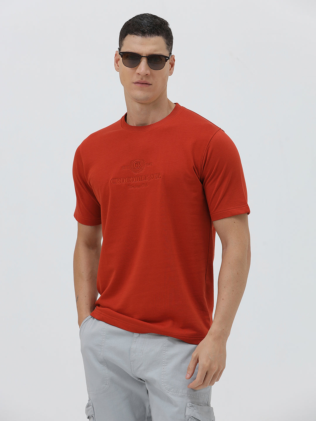 A man wearing a short-sleeve crewneck t-shirt in a muted brick red color, paired with light gray cargo pants, standing against a light gray studio background.
