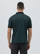 A man wearing a dark green short-sleeve polo shirt with a black collar and black sleeve trim, paired with dark gray jeans, against a light gray studio background.