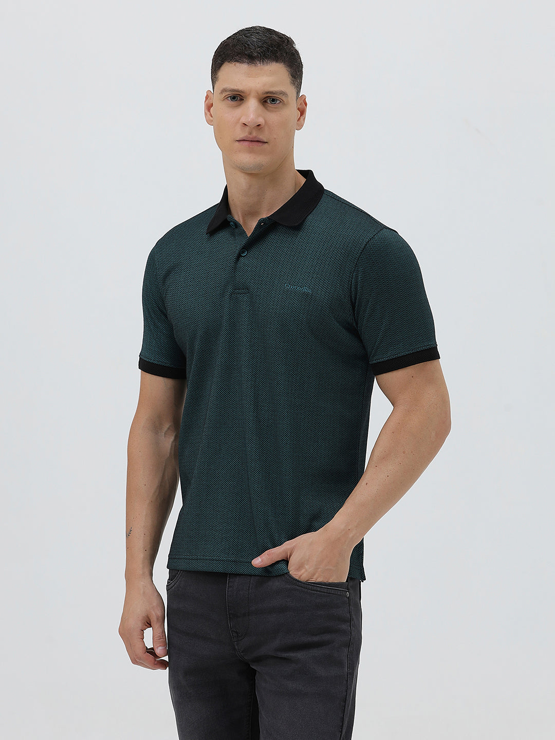 A man wearing a dark green short-sleeve polo shirt with a black collar and black sleeve trim, paired with dark gray jeans, against a light gray studio background.