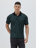 A man wearing a dark green short-sleeve polo shirt with a black collar and black sleeve trim, paired with dark gray jeans, against a light gray studio background.