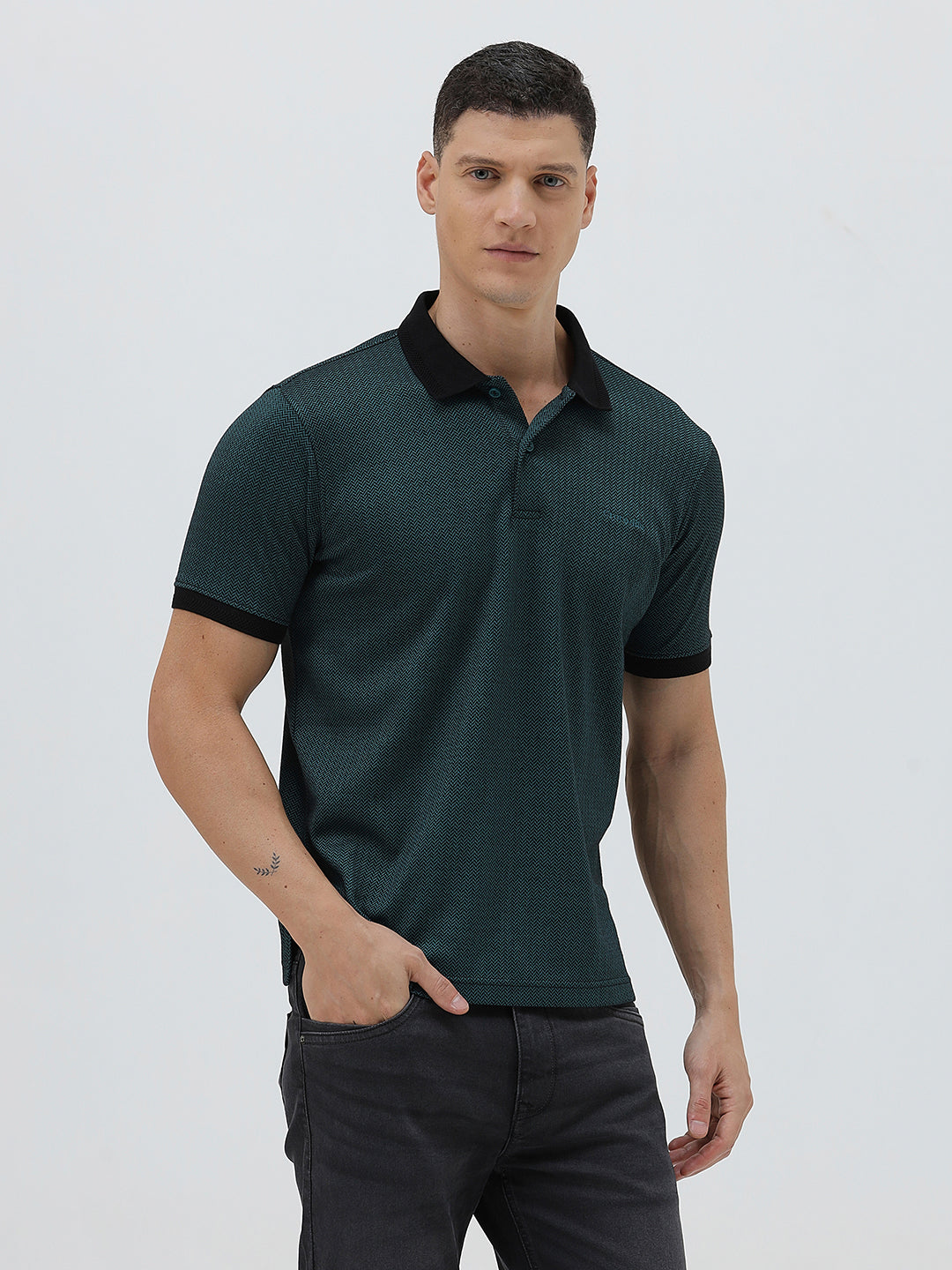 A man wearing a dark green short-sleeve polo shirt with a black collar and black sleeve trim, paired with dark gray jeans, against a light gray studio background.