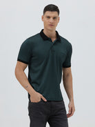A man wearing a dark green short-sleeve polo shirt with a black collar and black sleeve trim, paired with dark gray jeans, against a light gray studio background.