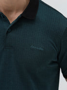A man wearing a dark green short-sleeve polo shirt with a black collar and black sleeve trim, paired with dark gray jeans, against a light gray studio background.