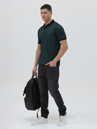 A man wearing a dark green short-sleeve polo shirt with a black collar and black sleeve trim, paired with dark gray jeans, against a light gray studio background.
