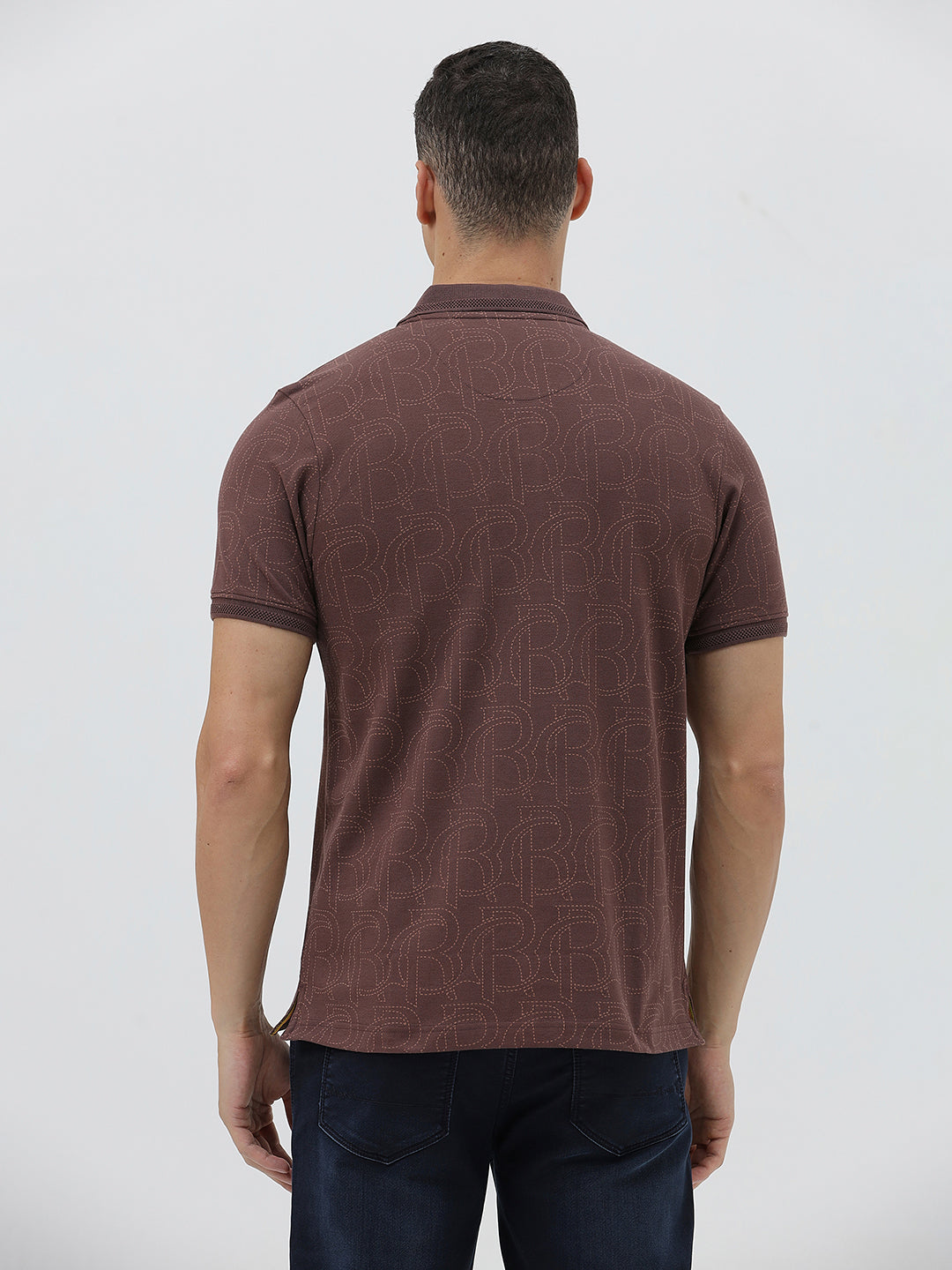 A man wearing a short-sleeve polo shirt in a dark brownish-rose color with a subtle tonal geometric pattern, paired with dark jeans, against a light gray studio background.