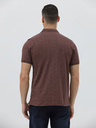 A man wearing a short-sleeve polo shirt in a dark brownish-rose color with a subtle tonal geometric pattern, paired with dark jeans, against a light gray studio background.