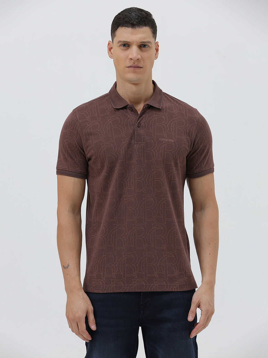 A man wearing a short-sleeve polo shirt in a dark brownish-rose color with a subtle tonal geometric pattern, paired with dark jeans, against a light gray studio background.