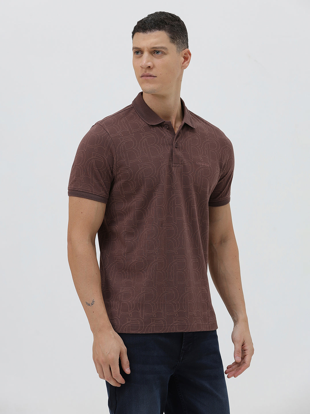 A man wearing a short-sleeve polo shirt in a dark brownish-rose color with a subtle tonal geometric pattern, paired with dark jeans, against a light gray studio background.