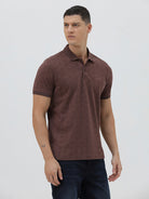 A man wearing a short-sleeve polo shirt in a dark brownish-rose color with a subtle tonal geometric pattern, paired with dark jeans, against a light gray studio background.