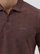 A man wearing a short-sleeve polo shirt in a dark brownish-rose color with a subtle tonal geometric pattern, paired with dark jeans, against a light gray studio background.