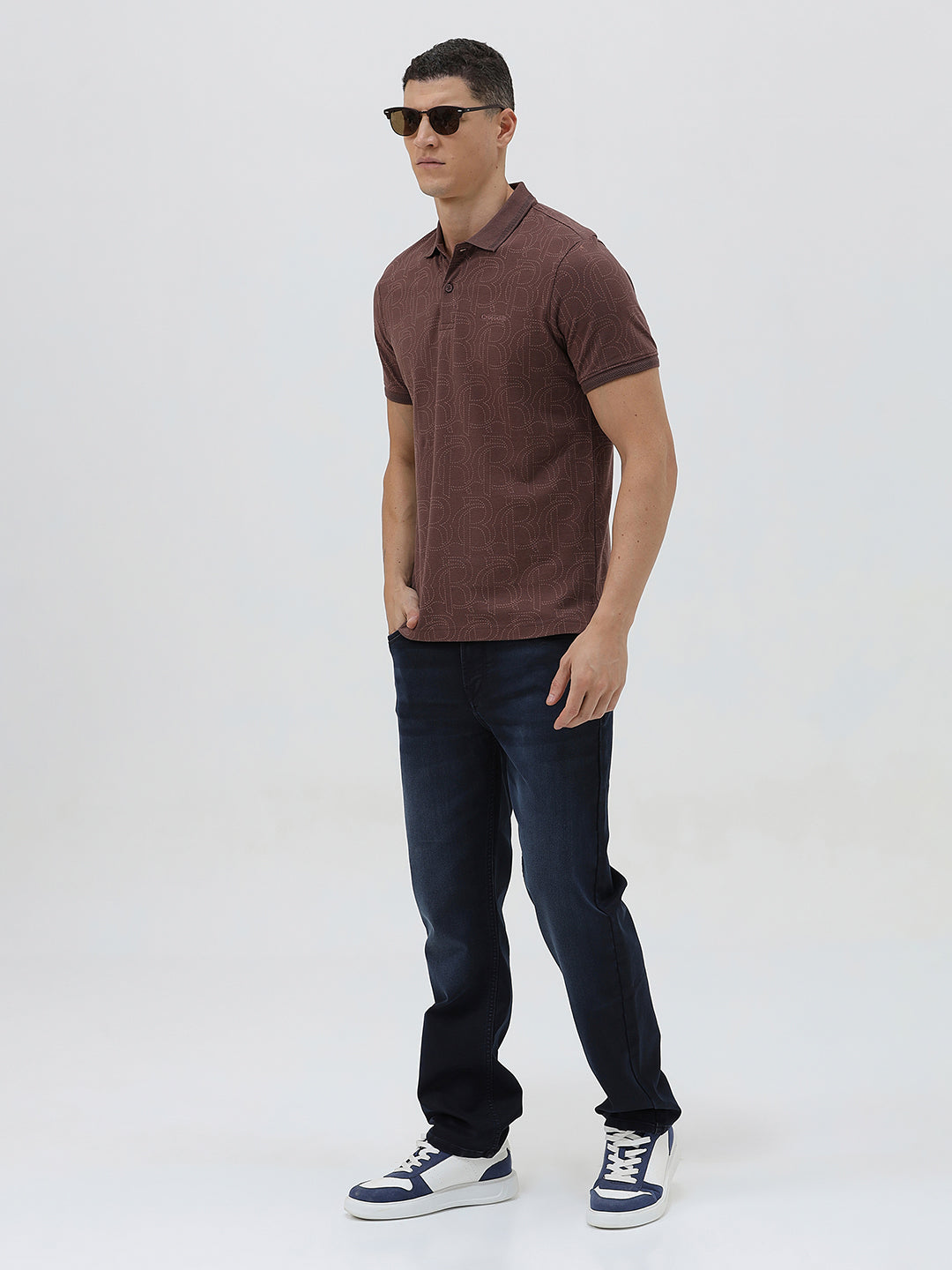 A man wearing a short-sleeve polo shirt in a dark brownish-rose color with a subtle tonal geometric pattern, paired with dark jeans, against a light gray studio background.