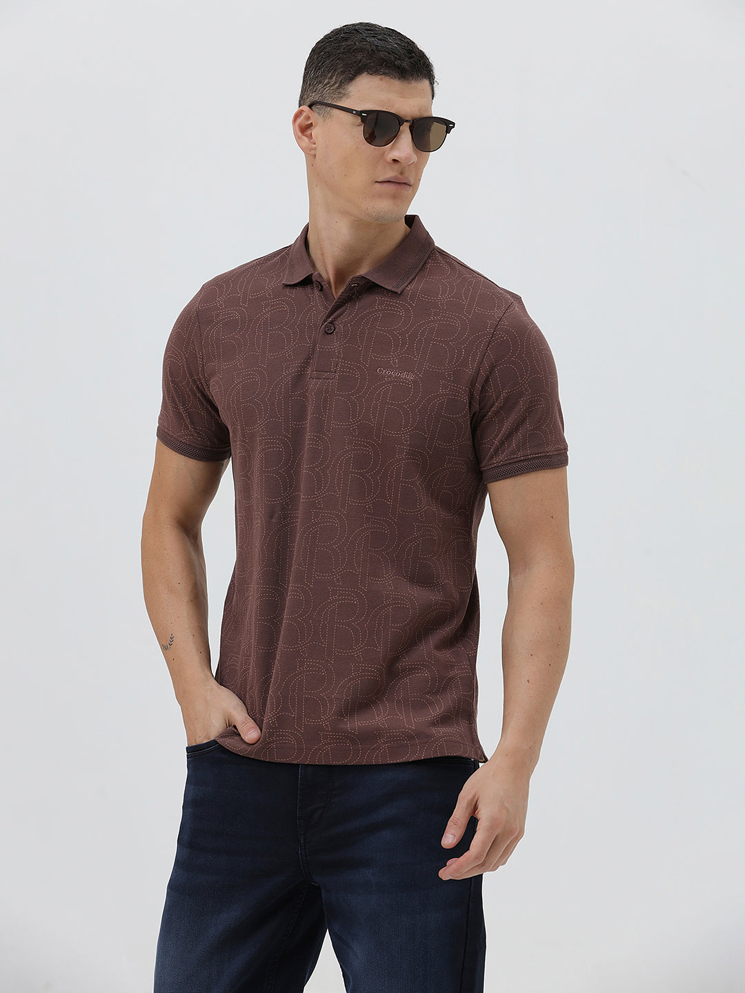 A man wearing a short-sleeve polo shirt in a dark brownish-rose color with a subtle tonal geometric pattern, paired with dark jeans, against a light gray studio background.