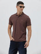 A man wearing a short-sleeve polo shirt in a dark brownish-rose color with a subtle tonal geometric pattern, paired with dark jeans, against a light gray studio background.