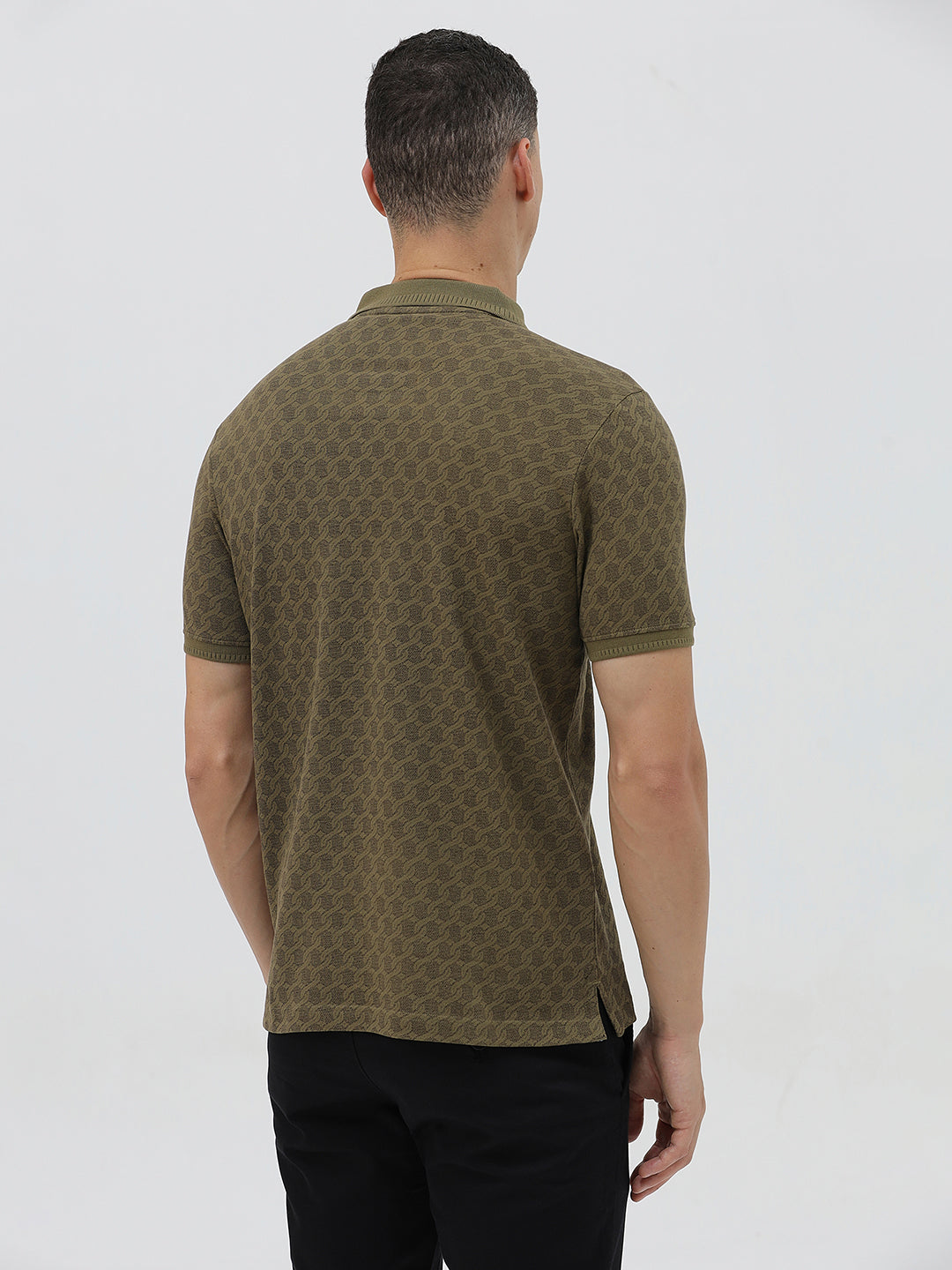 A man wearing a short-sleeve polo shirt with a subtle geometric monogram pattern in olive green and brown, paired with black trousers, against a light gray studio background.