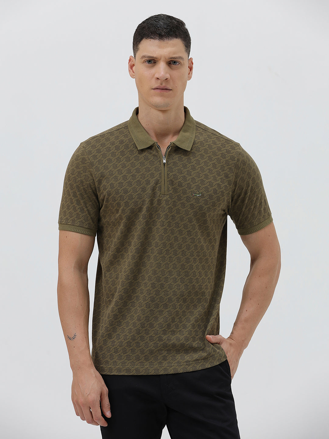 A man wearing a short-sleeve polo shirt with a subtle geometric monogram pattern in olive green and brown, paired with black trousers, against a light gray studio background.