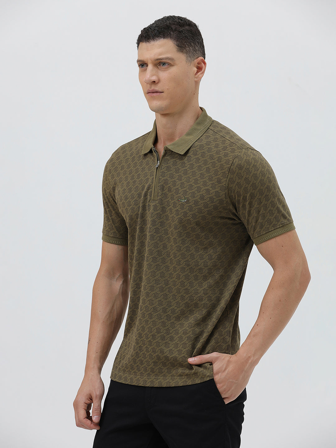 A man wearing a short-sleeve polo shirt with a subtle geometric monogram pattern in olive green and brown, paired with black trousers, against a light gray studio background.