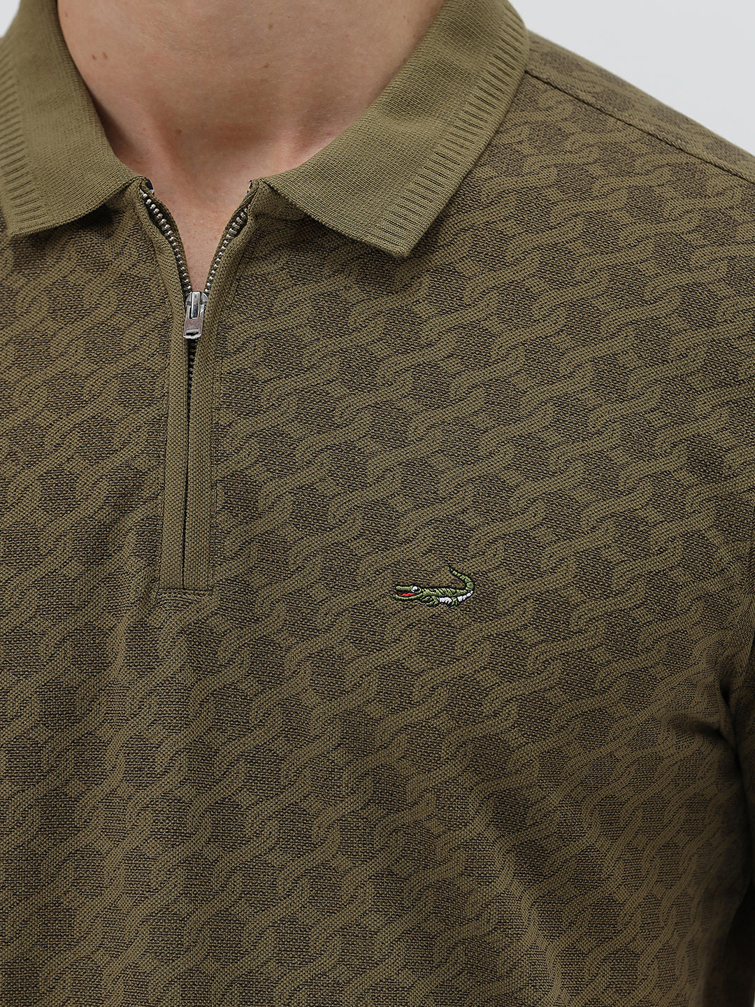 A man wearing a short-sleeve polo shirt with a subtle geometric monogram pattern in olive green and brown, paired with black trousers, against a light gray studio background.