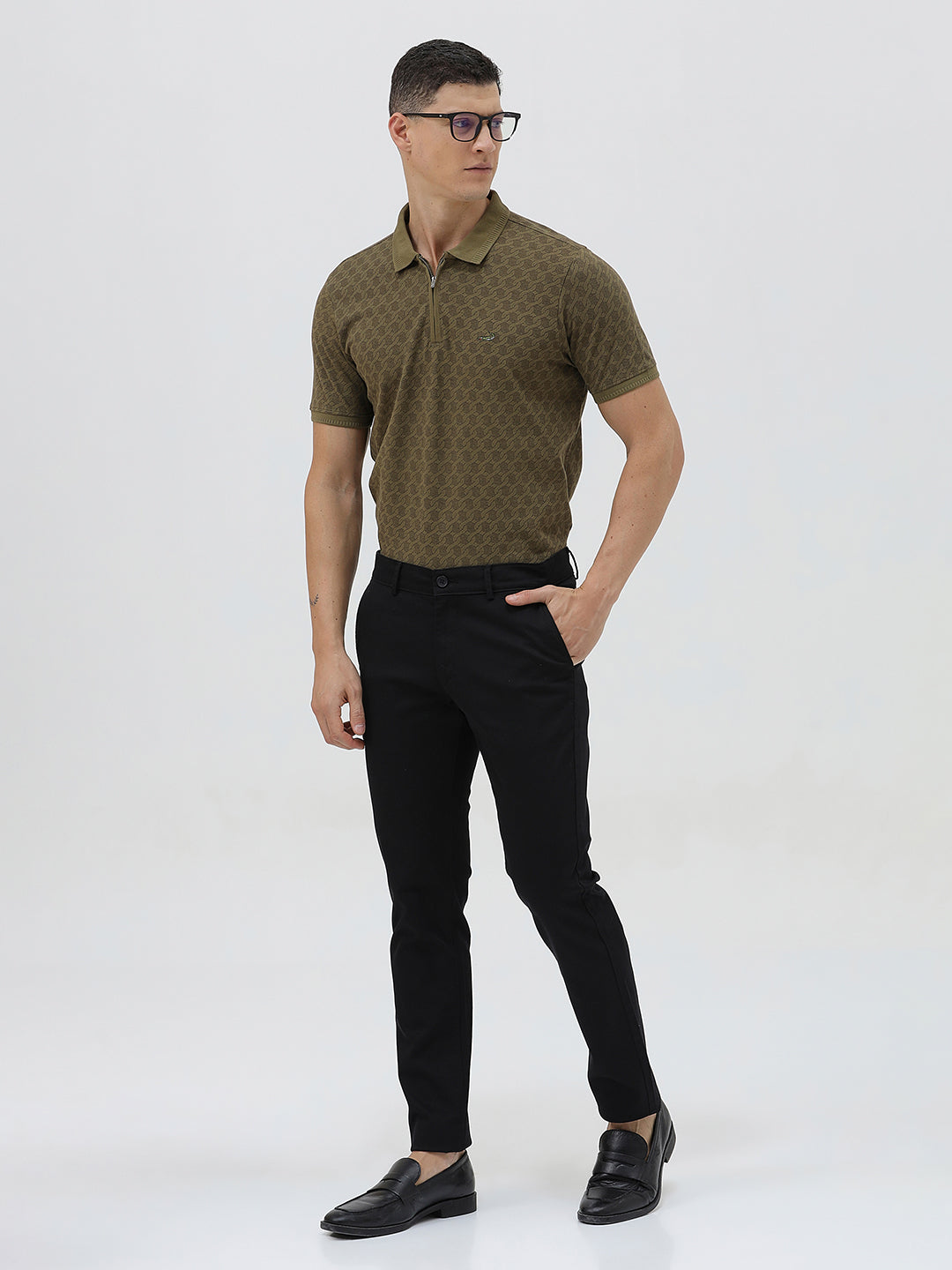 A man wearing a short-sleeve polo shirt with a subtle geometric monogram pattern in olive green and brown, paired with black trousers, against a light gray studio background.