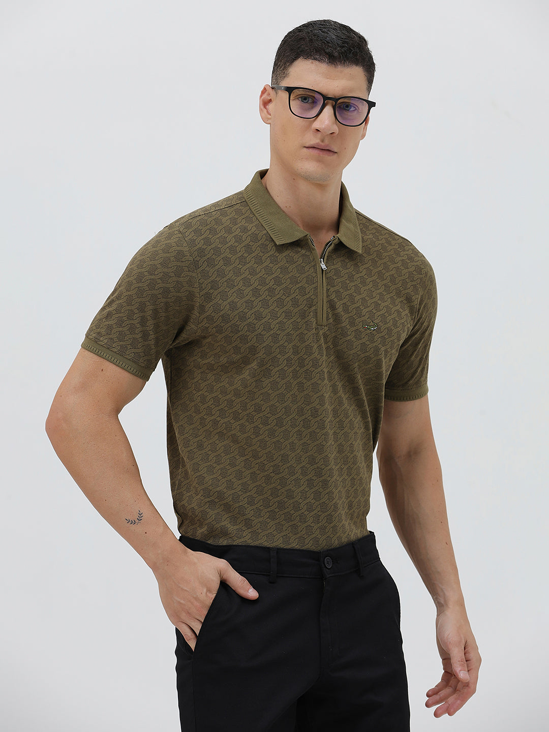 A man wearing a short-sleeve polo shirt with a subtle geometric monogram pattern in olive green and brown, paired with black trousers, against a light gray studio background.