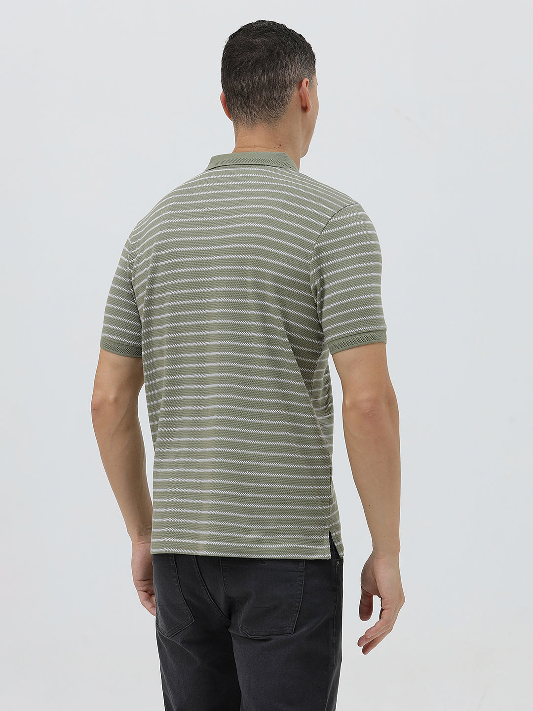 A man wearing a short-sleeve polo shirt with thin horizontal olive green and white stripes, paired with dark gray pants, standing against a light gray studio background.