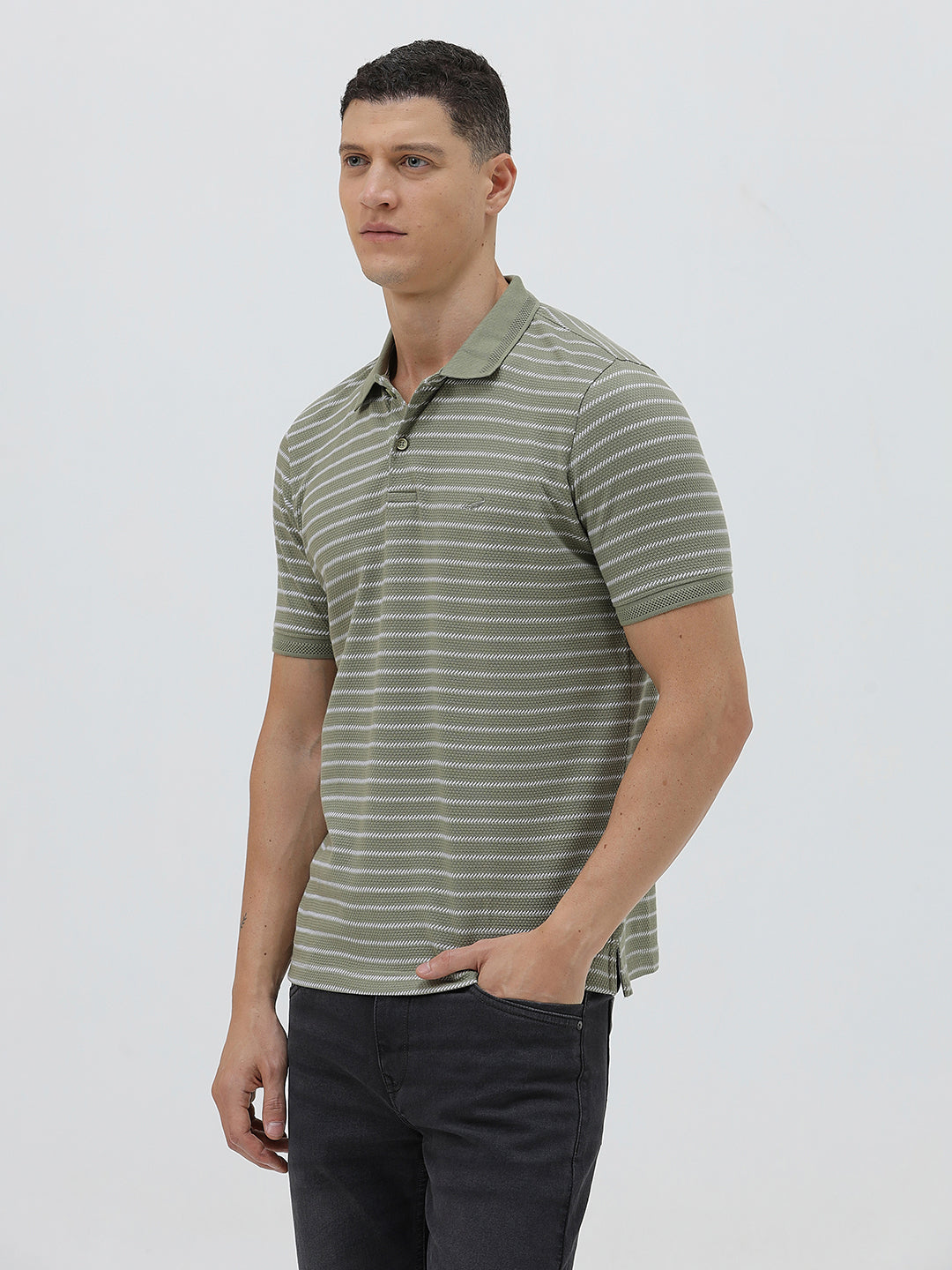 A man wearing a short-sleeve polo shirt with thin horizontal olive green and white stripes, paired with dark gray pants, standing against a light gray studio background.
