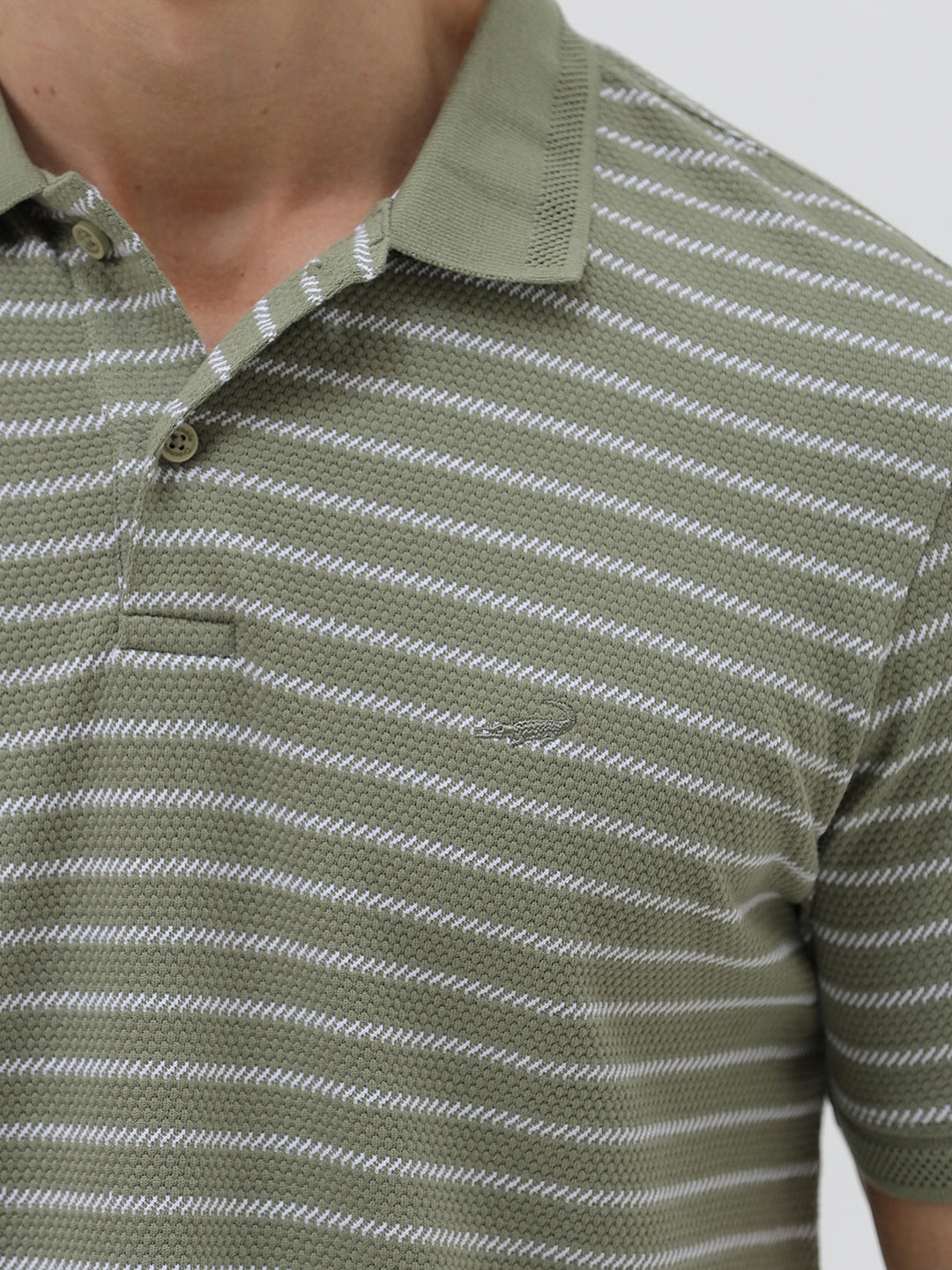A man wearing a short-sleeve polo shirt with thin horizontal olive green and white stripes, paired with dark gray pants, standing against a light gray studio background.