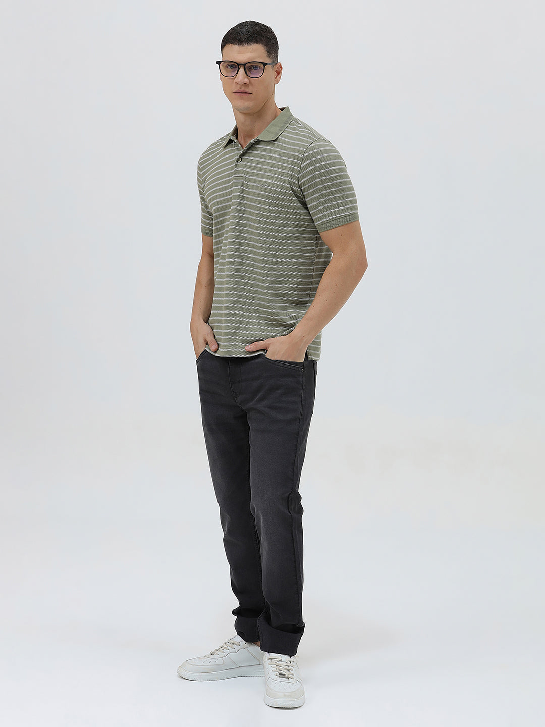 A man wearing a short-sleeve polo shirt with thin horizontal olive green and white stripes, paired with dark gray pants, standing against a light gray studio background.