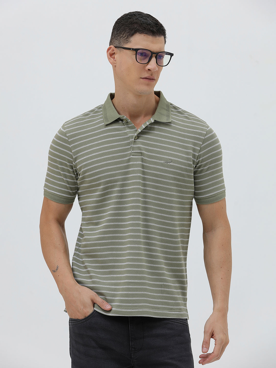 A man wearing a short-sleeve polo shirt with thin horizontal olive green and white stripes, paired with dark gray pants, standing against a light gray studio background.