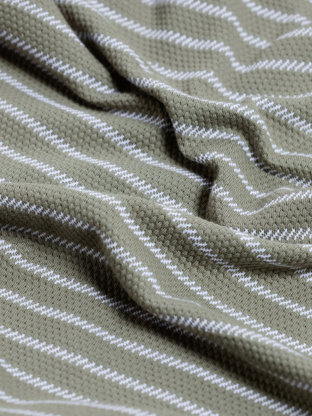 A man wearing a short-sleeve polo shirt with thin horizontal olive green and white stripes, paired with dark gray pants, standing against a light gray studio background.