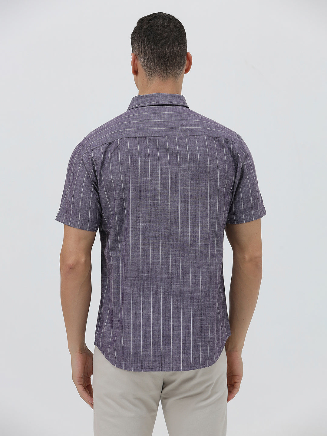 A man wearing a short-sleeve button-up shirt with a purple and white vertical stripe pattern, paired with light beige trousers, standing against a light gray studio background.