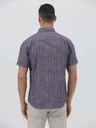 A man wearing a short-sleeve button-up shirt with a purple and white vertical stripe pattern, paired with light beige trousers, standing against a light gray studio background.