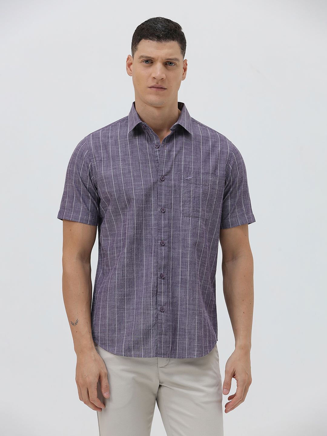 A man wearing a short-sleeve button-up shirt with a purple and white vertical stripe pattern, paired with light beige trousers, standing against a light gray studio background.