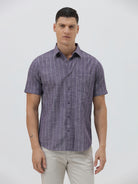 A man wearing a short-sleeve button-up shirt with a purple and white vertical stripe pattern, paired with light beige trousers, standing against a light gray studio background.