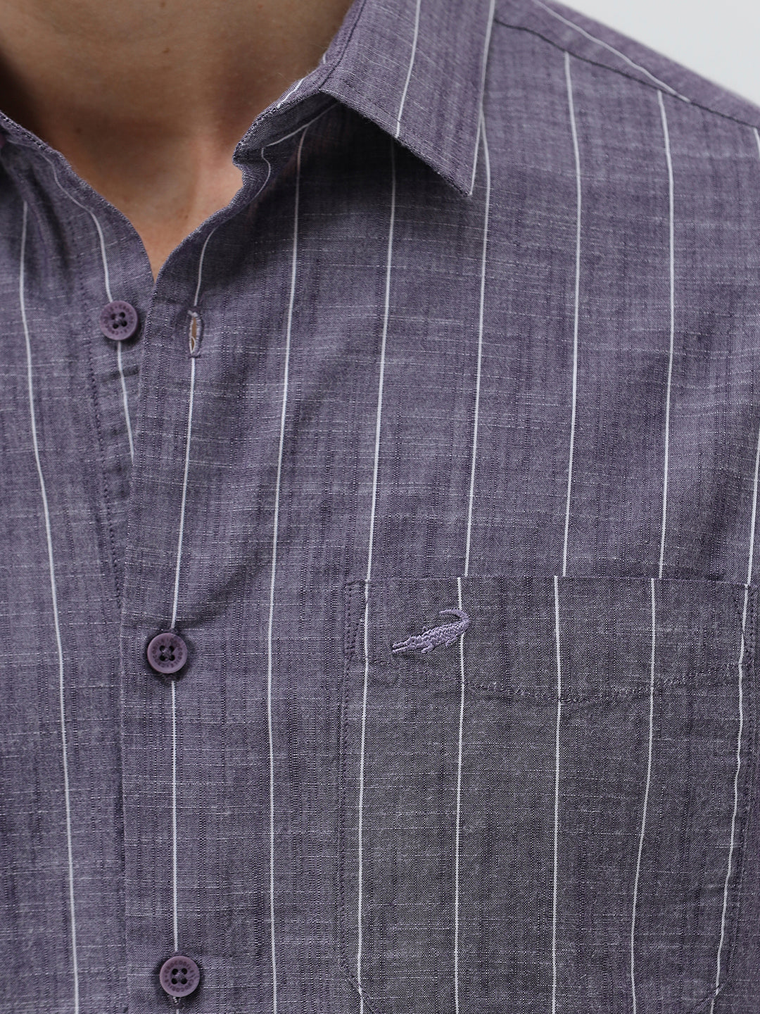 A man wearing a short-sleeve button-up shirt with a purple and white vertical stripe pattern, paired with light beige trousers, standing against a light gray studio background.