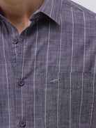 A man wearing a short-sleeve button-up shirt with a purple and white vertical stripe pattern, paired with light beige trousers, standing against a light gray studio background.