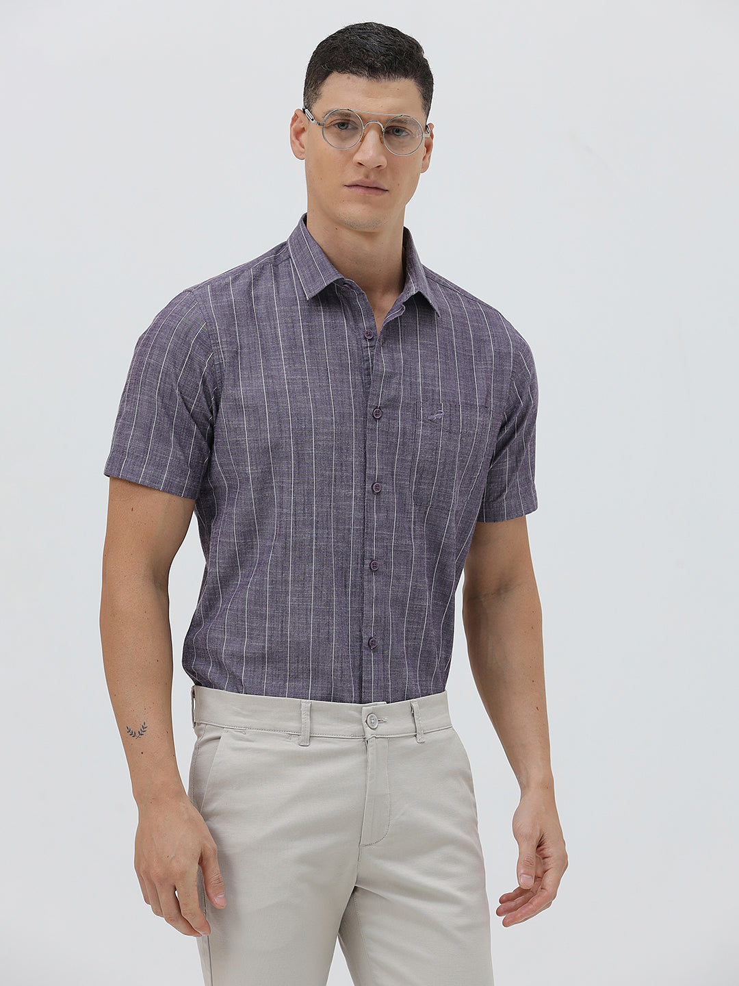 A man wearing a short-sleeve button-up shirt with a purple and white vertical stripe pattern, paired with light beige trousers, standing against a light gray studio background.