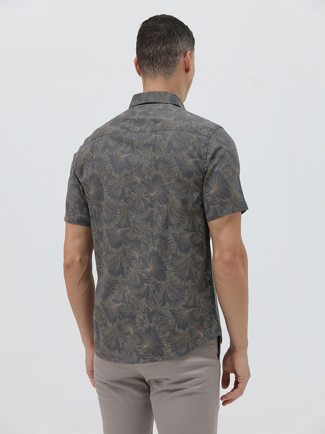 A man wearing a short-sleeve button-up shirt with a dark gray and brown palm leaf pattern, paired with light gray trousers, standing against a light gray background.