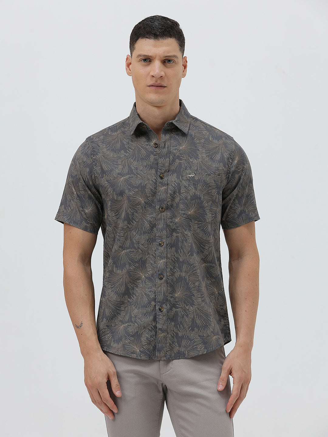 A man wearing a short-sleeve button-up shirt with a dark gray and brown palm leaf pattern, paired with light gray trousers, standing against a light gray background.