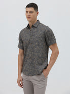 A man wearing a short-sleeve button-up shirt with a dark gray and brown palm leaf pattern, paired with light gray trousers, standing against a light gray background.
