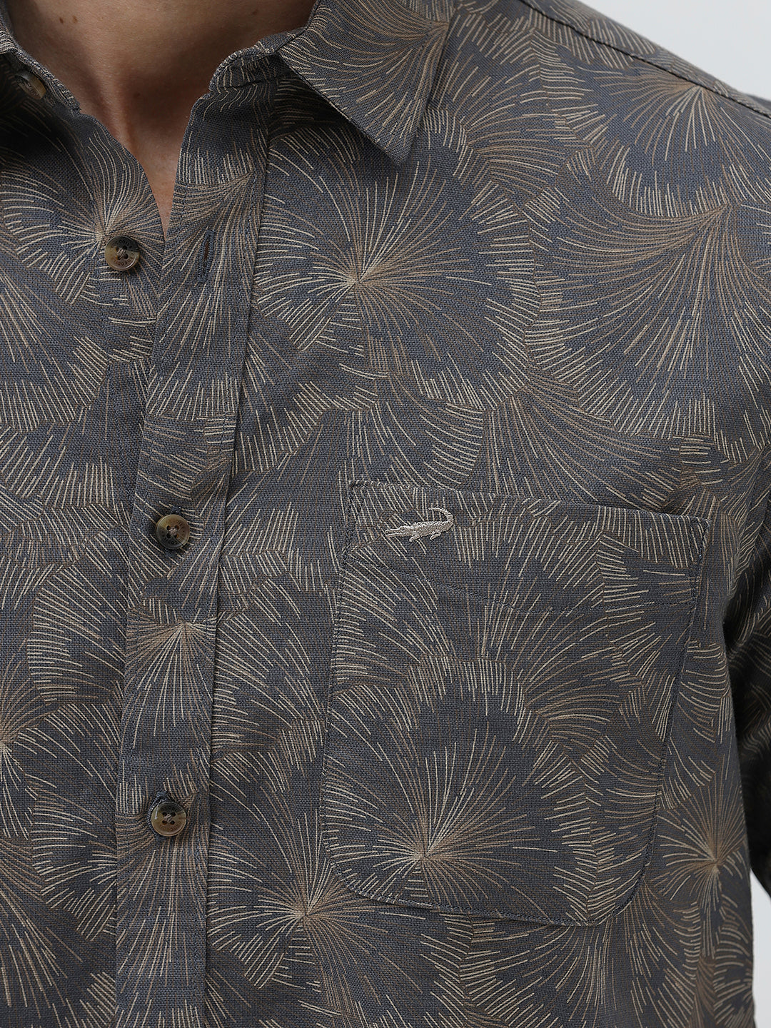 A man wearing a short-sleeve button-up shirt with a dark gray and brown palm leaf pattern, paired with light gray trousers, standing against a light gray background.