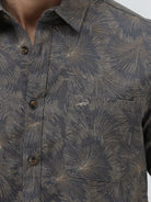 A man wearing a short-sleeve button-up shirt with a dark gray and brown palm leaf pattern, paired with light gray trousers, standing against a light gray background.