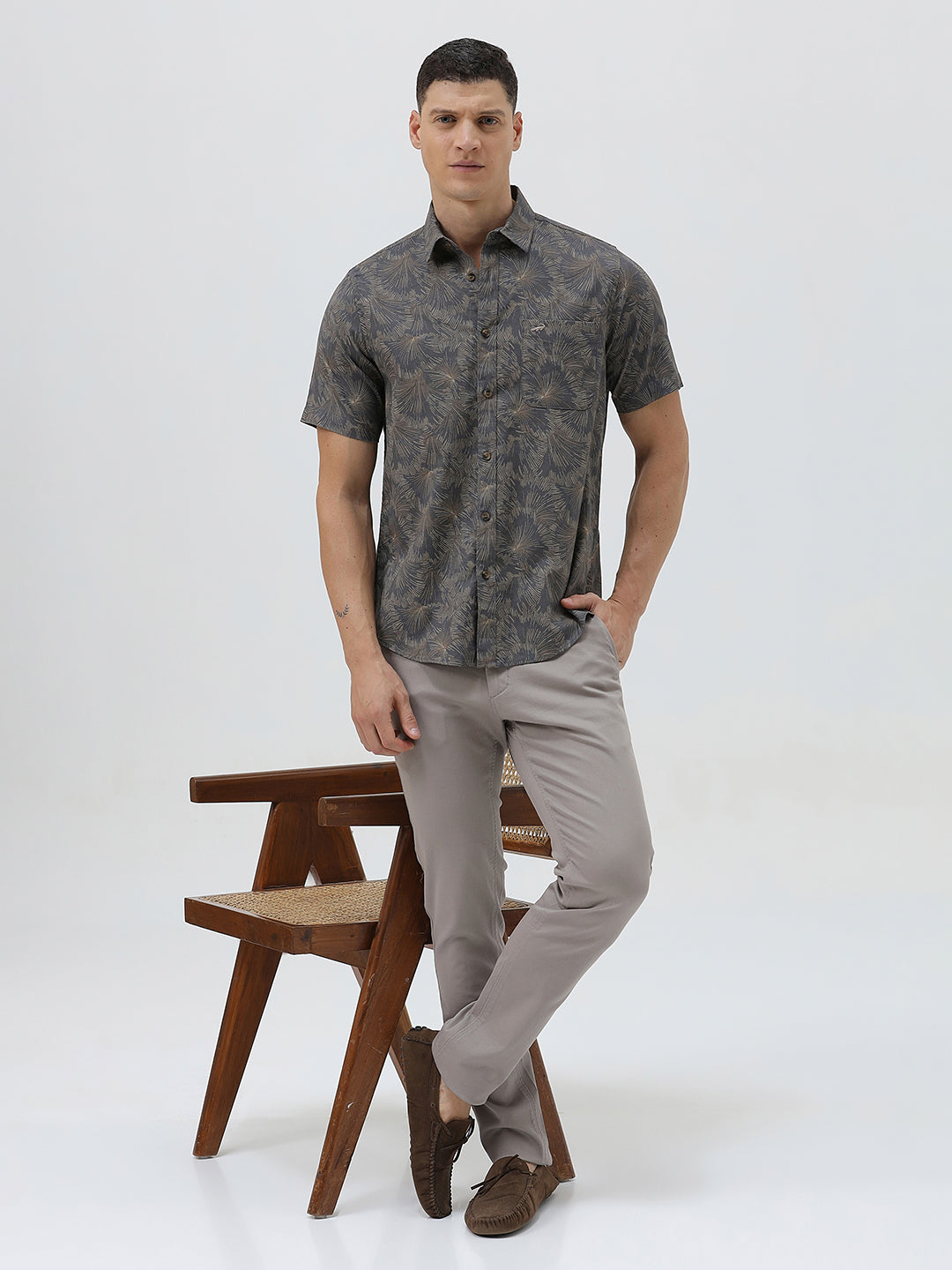 A man wearing a short-sleeve button-up shirt with a dark gray and brown palm leaf pattern, paired with light gray trousers, standing against a light gray background.