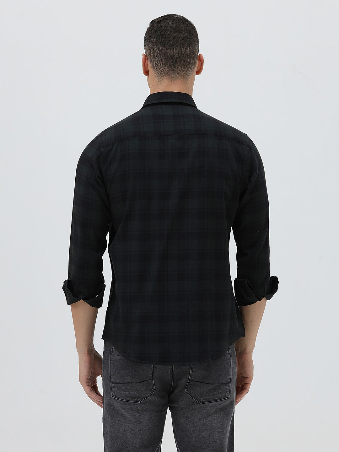A man wearing a black long-sleeve button-up shirt with a subtle dark plaid pattern, paired with dark gray jeans, standing against a light gray studio background.