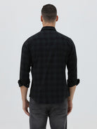 A man wearing a black long-sleeve button-up shirt with a subtle dark plaid pattern, paired with dark gray jeans, standing against a light gray studio background.